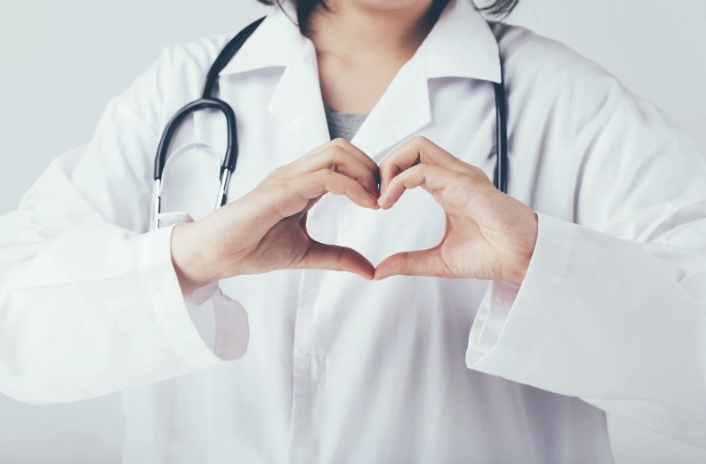Female wearing a lab gown with a stethoscope on her shoulder, forming a heart shape with her hand